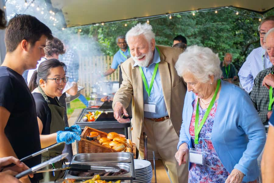 An elderly man at a food full of burgers and buns pointing out the food he wants to the staff serving him.