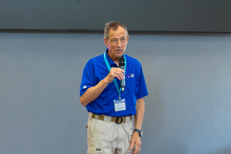 A middled aged man with a bright blue polo shirt speaking with a microphone at an event.