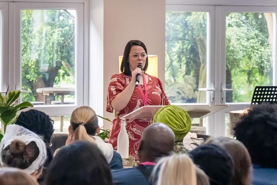 A woman in a red floral shirt speaking at an event with a microphone.