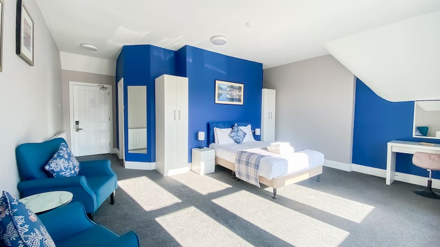 A bright, large bedroom with white and blue walls and light streaming through the windows showing a bed, a seating area, and other bedroom furniture such as a wardrobe and chest of drawers.