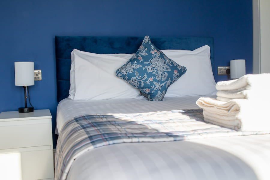 A bed with white sheets a blue cushion with a pattern which matches with the blue wall behind the bed.