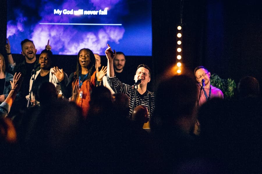 A man with his hand up worshipping in a dark room on stage with the worship team overlooking a crowd of people in the Derbyshire Hall at The Hayes Centre.