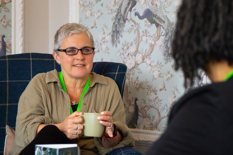 A woman holding a mug of coffee while listening to her friend in the Woodlands Lounge at High Leigh conference centre.