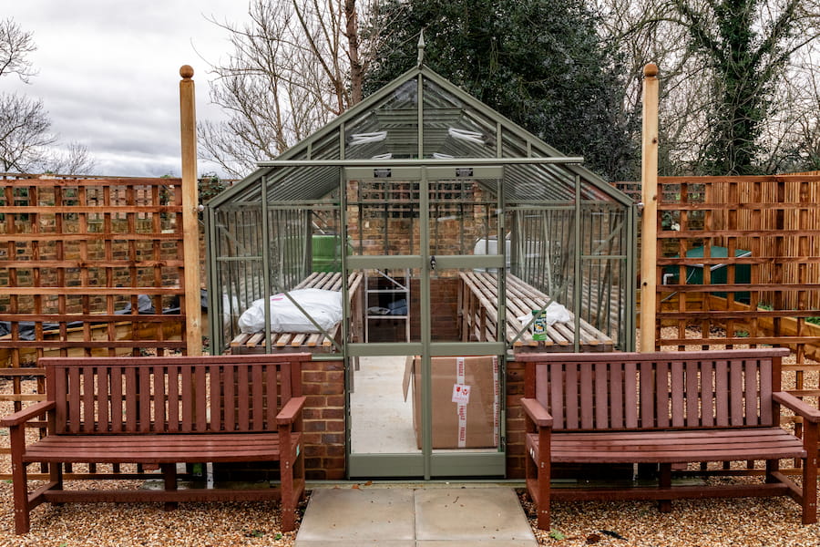 A closer image of the green house exterior with two garden benches either side of the entrance.