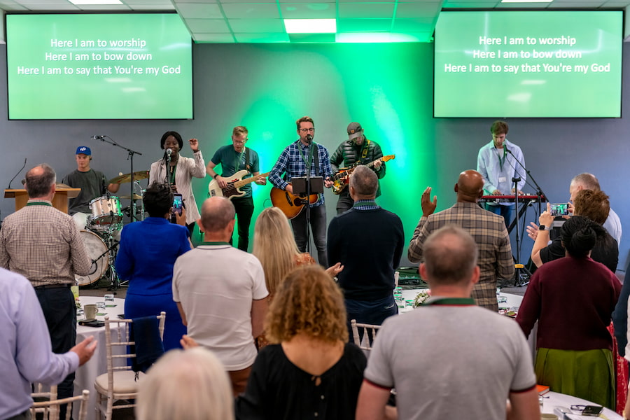 Lots of people worshipping with a view of the front stage showing green lights behind the band who are leading in worship.