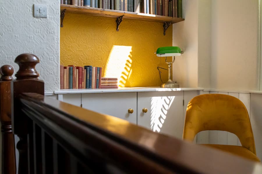 A chair in the corner of a cosy reading spot in front of a yellow wall which has a shelf with books and a small green reading lamp.
